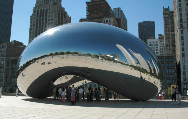 Large reflective sculpture (Cloud Gate or “The Bean”) in Chicago’s Millennium Park, with people gathered underneath and city skyline mirrored on its surface.
