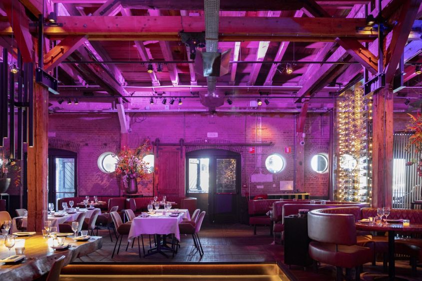Restaurant met bakstenen muren, paarse verlichting, houten balken en gedekte tafels.