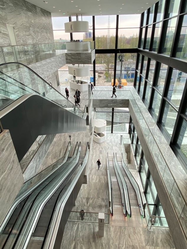 Moderne lobby met marmeren muren, glazen balustrades en roltrappen. G- en K-borden zichtbaar.