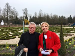 Man met bril en vrouw in rode jas poseren voor een tuin met buxushagen, bomen en een fontein.