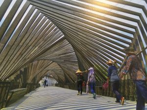 Houten overkapping met schaduwstrepen, mensen lopen op pad, gebouwen in de verte.
