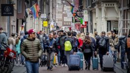 Mensen met koffers lopen op drukke straat met winkels en regenboogvlaggen.