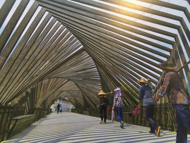 Houten overkapping met schaduwstrepen, mensen lopen op pad, gebouwen in de verte.