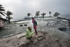 Twee vrouwen in traditionele kleding staan op een modderige oever bij palmbomen, met op de achtergrond gebouwen en water.