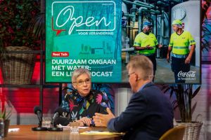 Een vrouw zit aan tafel, praat met een man. Op scherm: "Open voor verandering".