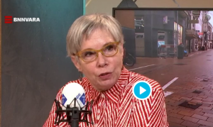 Vrouw met bril en rood-wit gestreepte blouse spreekt, met BNNVARA logo en straatbeeld op scherm.