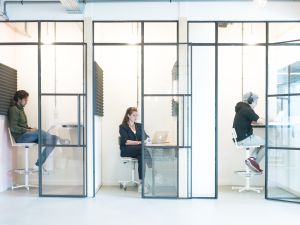 Drie personen werken in aparte glazen kantoorunits met laptops.