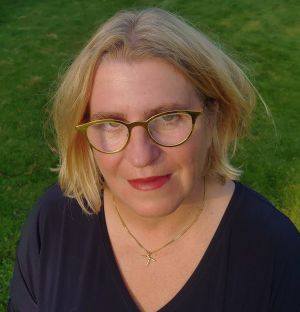 Een vrouw met blond haar en een gouden bril draagt een donkerblauw shirt en een ketting met een pickaxe-hanger.