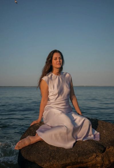 Een vrouw met lang donker haar en blote voeten zit op een rots aan de kust.