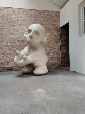 Een groot, wit, abstract beeld van een mensachtige figuur zit op een grijze betonnen vloer voor een bakstenen muur.