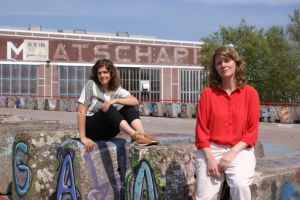 Twee vrouwen zitten op betonnen blokken voor een gebouw met de tekst "MATSCHAPPIJ" in grote letters.