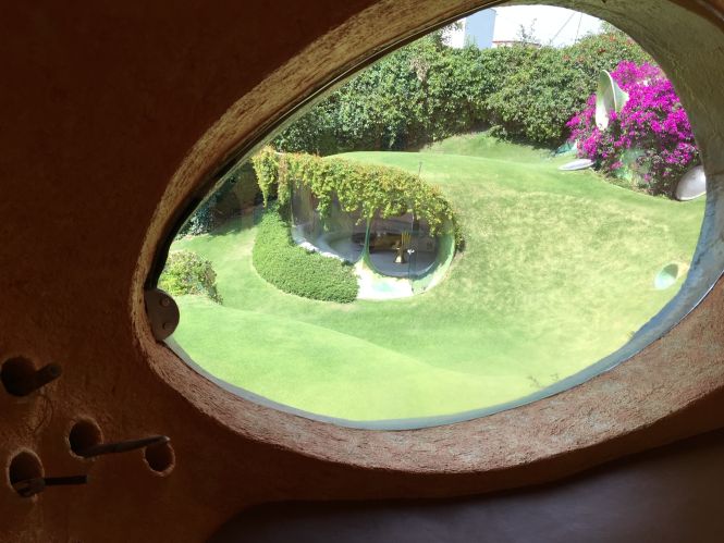Gouden hand en roze bloemen zichtbaar door een raam met organische vorm in een groene tuin.