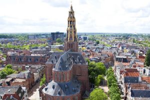 Kerk met toren in een stad met veel huizen en bomen, met op de achtergrond een donker gebouw.