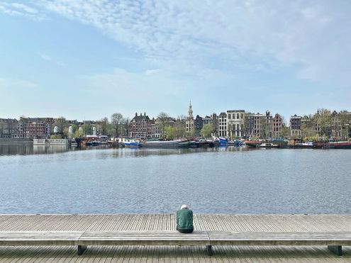 Persoon op bank kijkt uit over water naar Amsterdamse huizenrij met boten.