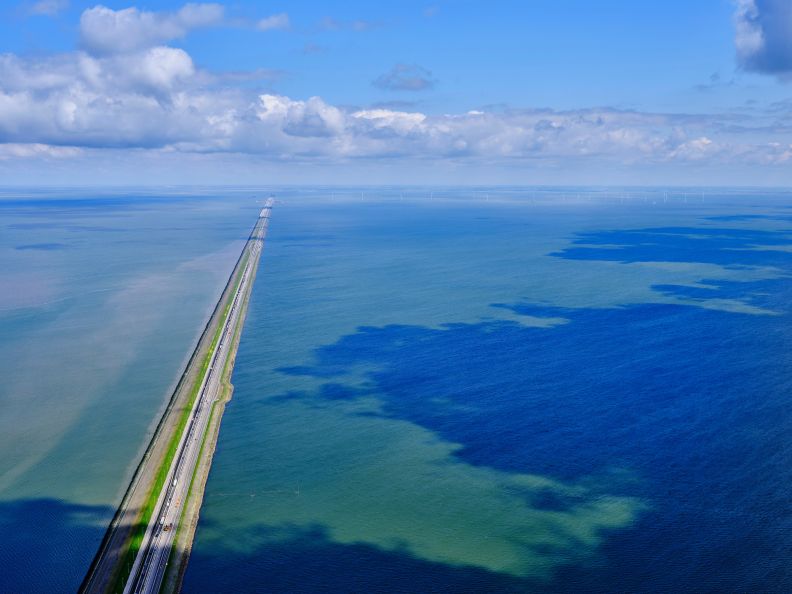 Lange dam met verkeer op het water met windturbines in de verte onder bewolkte blauwe hemel.