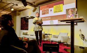 Vrouw spreekt op podium voor projectie "STADS LEVEN TRACY METZ EMPOWER THE CITY" met een lachende vrouw erop.