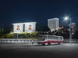 Bussen en portretten van leiders in Pyongyang bij nacht, met een bus op de voorgrond en twee grote portretten op een heuvel achtergrond.