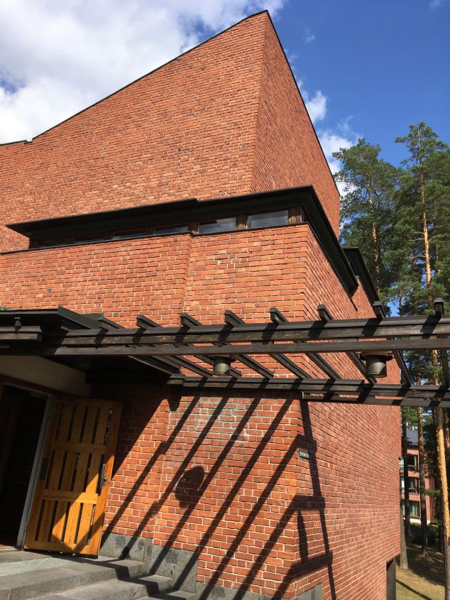 Bakstenen gebouw met houten pergola, trap en een open deur met daarboven kleine ramen.