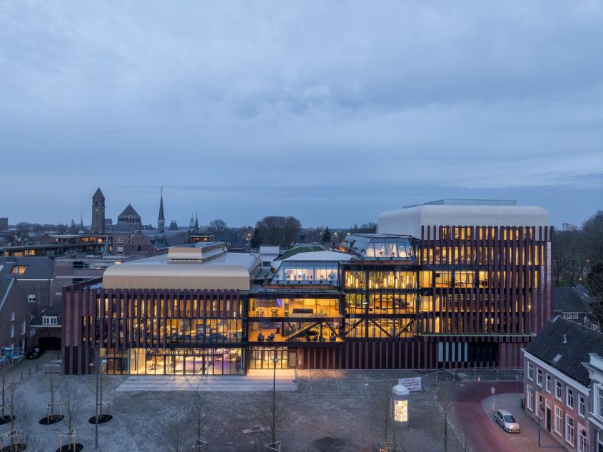 Modern gebouw met veel glas en baksteen, verlicht in de schemering.