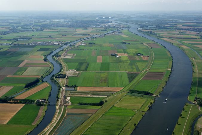 Luchtfoto van een brede rivier die door groen akkerland kronkelt, met een vrachtschip erop.