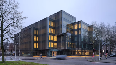 Moderne kantoorgebouwen met veel glas en stalen lamellen, 's avonds verlicht, met een mens die uit een raam kijkt.