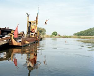 Jongen springt van gele boot in water, hond kijkt toe, zwemmer in de verte.