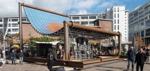 Constructie met grote gebogen zonnepanelen boven een openluchtruimte met mensen en gebouwen.