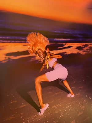 Vrouw met oranje pruik, houdt waterpistool vast, op strand bij zonsondergang.