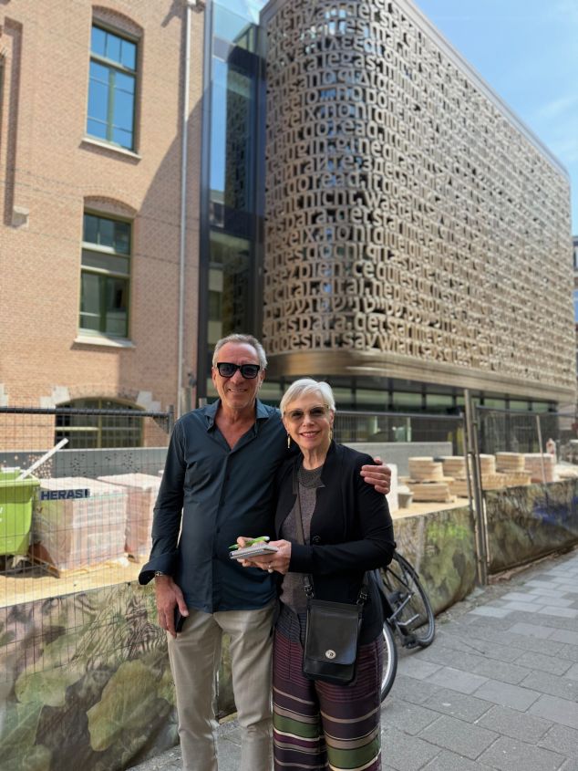 Man en vrouw glimlachen, vrouw houdt notitieboekje vast, gebouw met tekst.