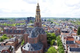 Kerk met toren in een stad met veel huizen en bomen, met op de achtergrond een donker gebouw.