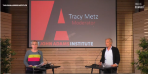 Drie mensen spreken op een podium met een John Adams Institute logo.