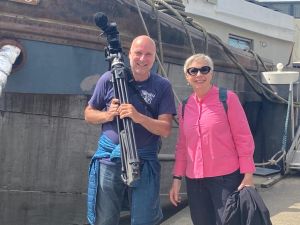 Man met videocamera en vrouw in roze shirt staan bij een boot