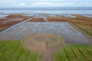 Megalithische cirkel van staande stenen in een vlak, modderig landschap bij eb, met de zee op de achtergrond.