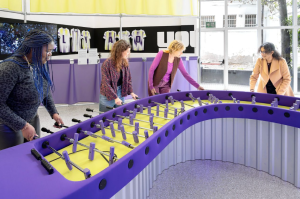 Foosball spel met vier vrouwen, gele en paarse elementen, muurschildering met "LA", "Y", "OU".