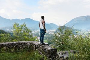 Man in wit tanktop en blauwe jeans staat op stenen muur, kijkt uit over vallei met dorp en bergen.