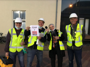 Vier personen, inclusief twee mannen met gele hesjes en helmen, met een bord "TEAM 7 HOOG WATER BRIGADE".