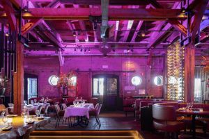 Restaurant met bakstenen muren, paarse verlichting, houten balken en gedekte tafels.