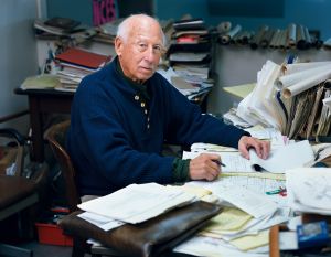 Een man met grijs haar en een blauwe trui zit aan een bureau vol papierwerk en schrijft op een notitieblok.