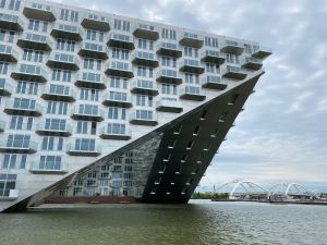Modern gebouw met glazen balkons aan het water, met een brug op de achtergrond.