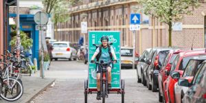 Bezorgpersoon op bakfiets met groene reclame met tekst "LID VAN FIETSERS.NL", rijdend op straat