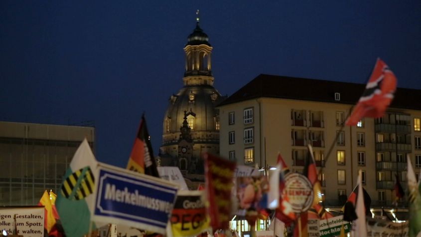 Vlaggen in de nacht voor verlichte gebouwen en een koepelkerk, met tekst die "Merkel muss weg" en "PEGIDA" aangeeft.
