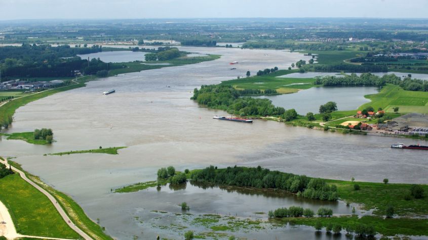 Vloedgolf met schepen op een brede rivier omgeven door groene oevers en weilanden.