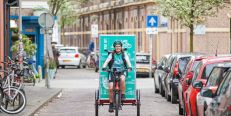 Bezorgpersoon op bakfiets met groene reclame met tekst "LID VAN FIETSERS.NL", rijdend op straat