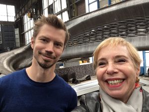 Man en vrouw maken een selfie voor een groot, grijs, 3D-geprint object in een industriële omgeving.