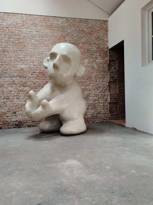 Een groot, wit, abstract beeld van een mensachtige figuur zit op een grijze betonnen vloer voor een bakstenen muur.
