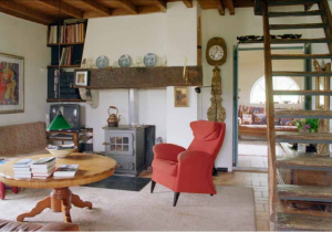 Zitkamer met rode fauteuil, houtkachel, ronde tafel met boeken en trap naar boven.