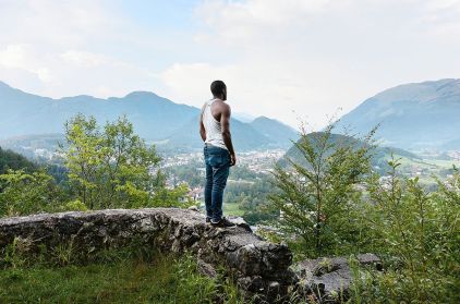 Man in wit tanktop en blauwe jeans staat op stenen muur, kijkt uit over vallei met dorp en bergen.