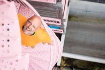 Kale man met oranje trui knipoogt op roze wenteltrap, met betonnen ondergrond ernaast.