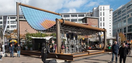 Constructie met grote gebogen zonnepanelen boven een openluchtruimte met mensen en gebouwen.