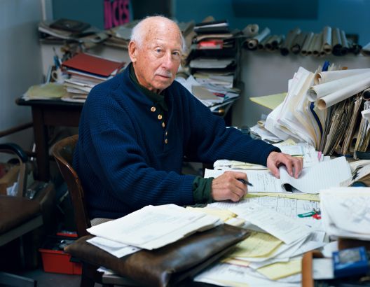 Een man met grijs haar en een blauwe trui zit aan een bureau vol papierwerk en schrijft op een notitieblok.
